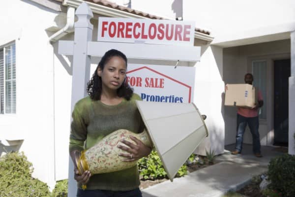 Distressed woman standing next to a 'Foreclosure' sign in her front yard.